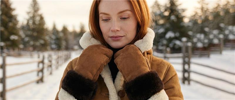Woman wearing brown shearling mittens and a tan sheepskin coat with a cream fur collar in a snowy winter setting.